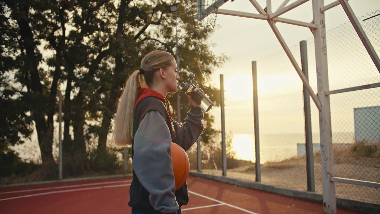 Close-up a blonde girl drinks water from a sports bottle and holds an orange basketball in her hands during her workout in the morning at Sunrise in the summer