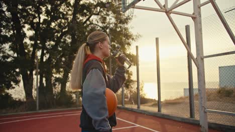 close-up una chica rubia bebe agua de una botella deportiva y sostiene una pelota de baloncesto naranja en sus manos durante su entrenamiento por la mañana al amanecer en el verano