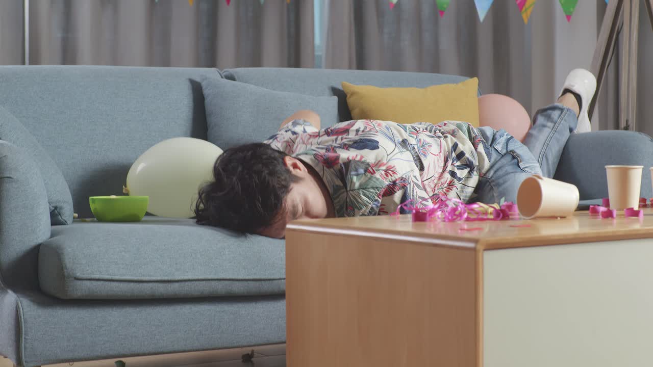 술에 취한 아시아 남자가 소파에서 잠을 자고 집에 있는 파티 에서 두통으로 인해 머리를 손으로 잡고 깨어난다.