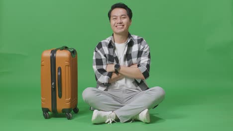 full body of asian male traveler with luggage and passport crossing arms and smiling to camera while sitting in the green screen background studio