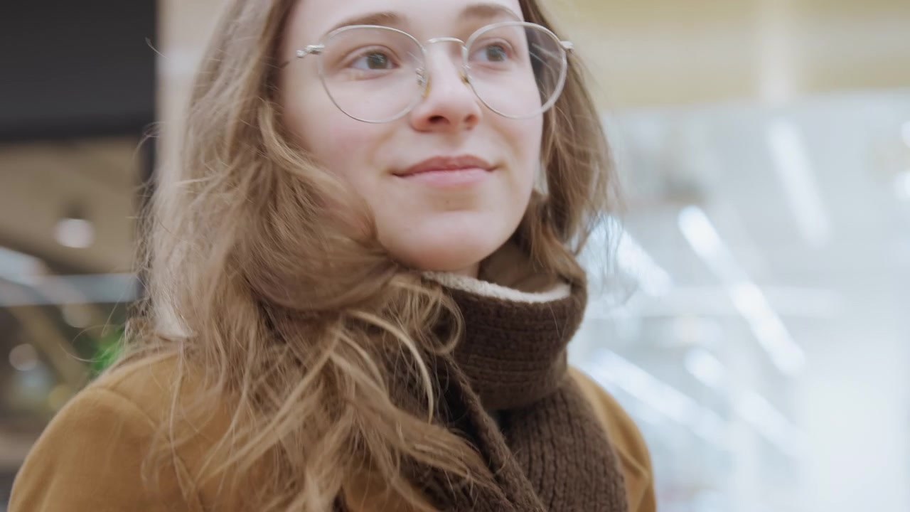 Jovem mulher sorrindo no shopping com casaco marrom e cachecol, retrato espontâneo em close-up com óculos e luzes de teto suaves, olhar gentil e expressão calorosa enquanto olha vitrines sob interior moderno.