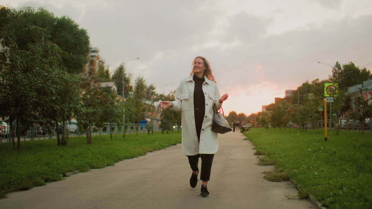 young girl walking along paved walkway twirling white umbrella happily while smiling during sunset with green grass, trees, distant people and signpost in background