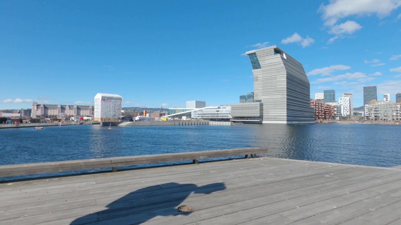 Vista ampla da orla de Oslo com horizonte moderno e porto, vista de um calçadão de madeira em um dia ensolarado.