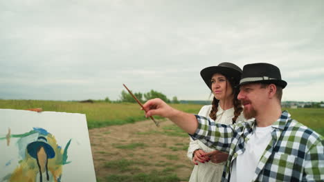 a drawer in a checkered shirt and hat points with a brush, pointing. a woman beside him, in a white dress and hat. they stand in a grassy field, with a partially painted canvas in the background