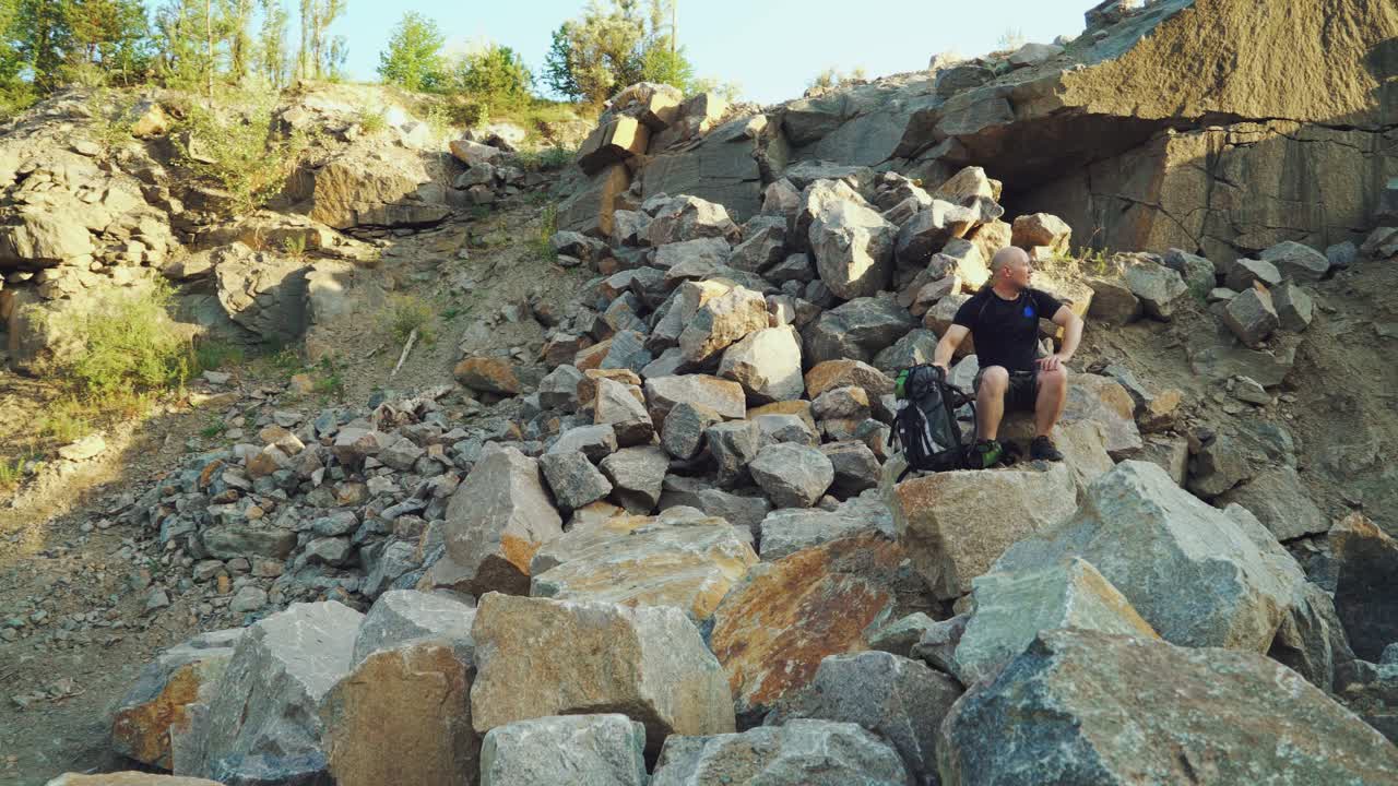 a traveler with a backpack in shorts and a black t-shirt is sitting on a large rock and examining a beautiful landscape around himself