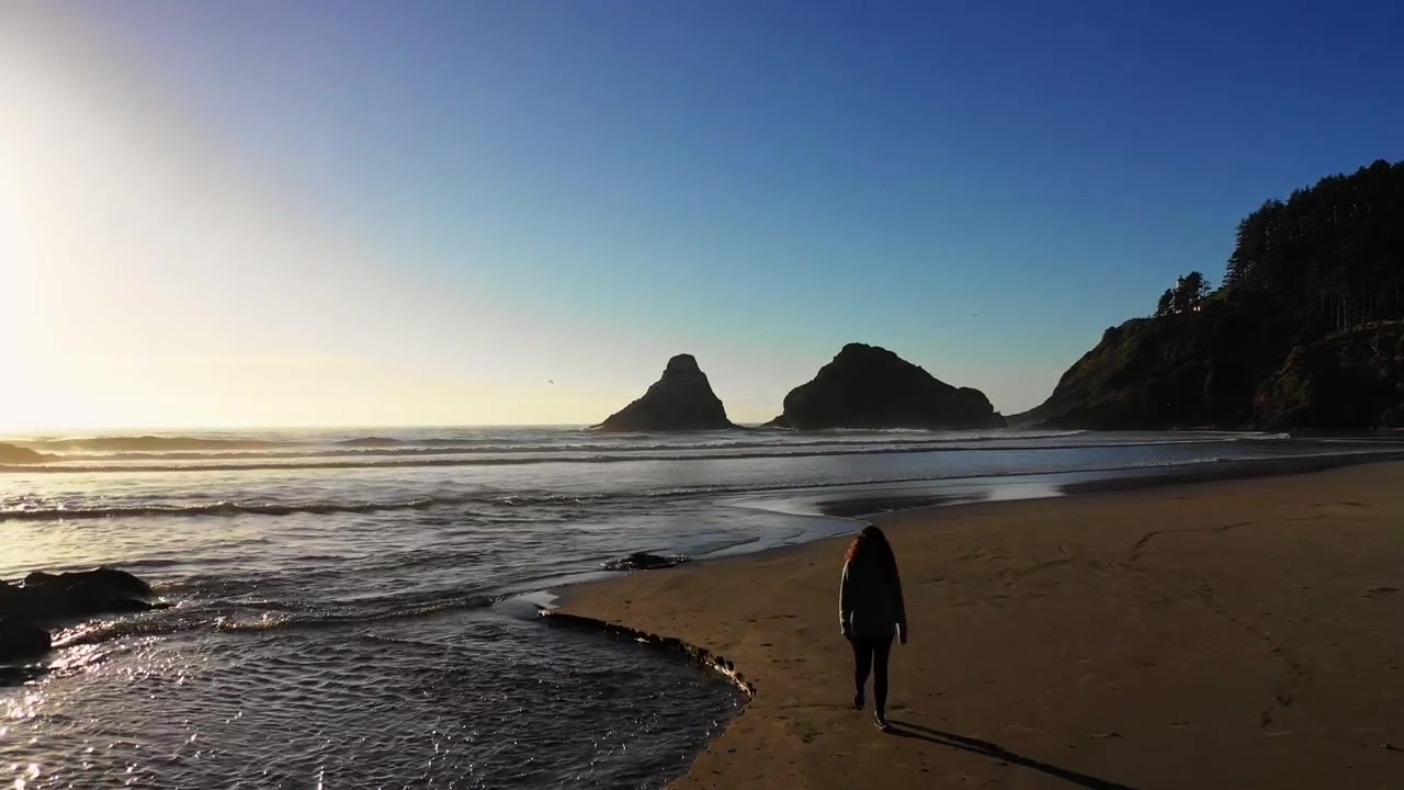 Una donna che si allontana dall'acqua mentre onde calme dell'Oceano Pacifico settentrionale si infrangono lungo la spiaggia sulla costa dell'Oregon al tramonto.