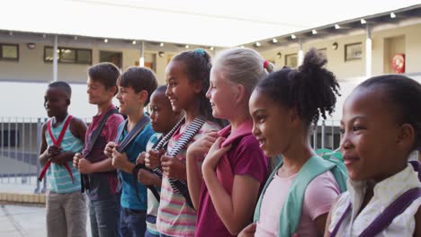 diverse group of schoolchildren wearing backpacks smiling and standing in a row at school yard