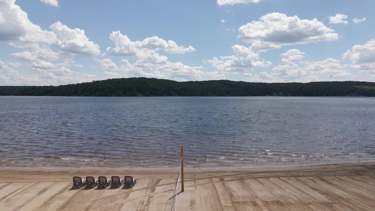 Sandstrand med stoler og volleyballnett, rolig strandlinje ved Commanda Lake i Restoule, Ontario, under en lys sommerhimmel, etablerende droneopptak.