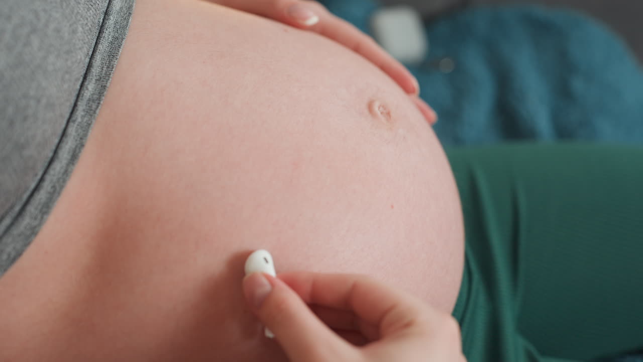 Primo piano di donna caucasica incinta che controlla un piccolo monitor sulla pancia, ambiente tranquillo del soggiorno, mano ferma che posiziona il sensore vicino all'ombelico, momento tranquillo di legame materno e rassicurazione.