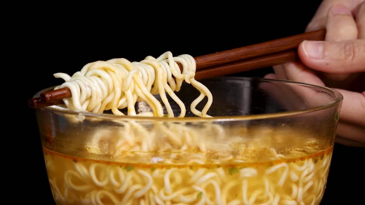 A hand uses chopsticks to lift and roll cooked instant noodles from a clear glass bowl filled with broth, under bright studio lighting and black background