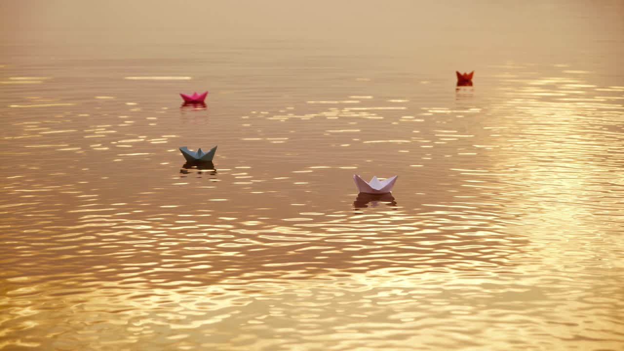 Colored paper ships floating in the river and some water splashes cover them a little at sunset. Four origami boat swim in the water