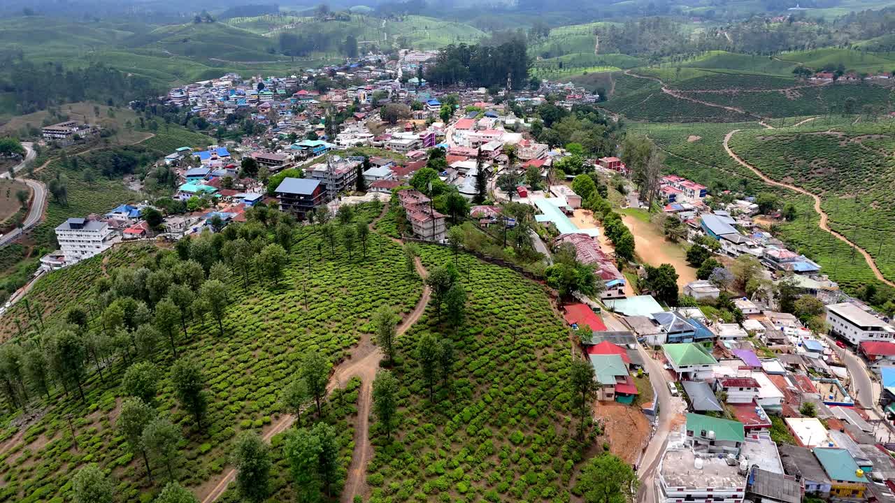Vista aérea cinematográfica de Valparai, exibindo uma vibrante cidade montanhosa com telhados coloridos espalhados ao longo de uma crista, cercada por exuberantes plantações de chá e estradas sinuosas através de colinas verdes ondulantes