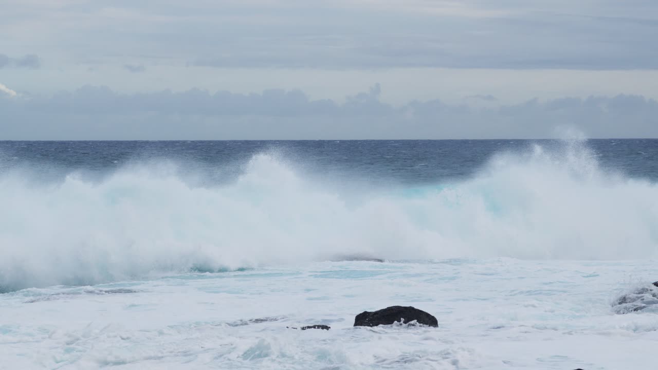 A teal wall of surf rears and folds near black shoreline rocks, wind slicing spray from the crest as foamy water surges landward beneath gray, unsettled skies
