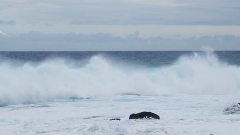 a teal wall of surf rears and folds near black shoreline rocks, wind slicing spray from the crest as foamy water surges landward beneath gray, unsettled skies
