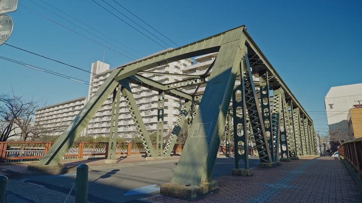 La travatura reticolare in acciaio verde del Ponte Fukuju domina l'inquadratura dal livello della strada a Tokyo, con marciapiede, alberi spogli e un grande complesso residenziale visibile dietro, ripreso come un'inquadratura statica grandangolare