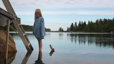 sad young blonde caucasian girl standing in front of lake water, tilt down