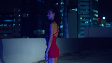 A-rooftop-at-night-hosts-a-young-girl-in-a-red-dress,-with-city-buildings-rising-in-the-background