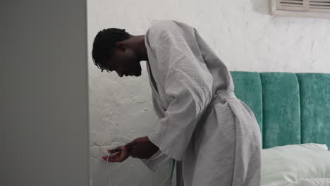 youth in bathrobe bending down by bed adjusting cords. cozy bedroom with green velvet headboard and soft bedding. quiet, intimate morning scene with focus on daily routine and self-care