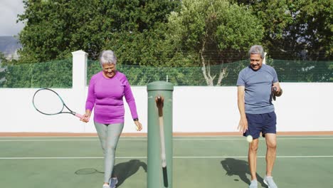 Video-Eines-Glücklichen-Gemischtrassigen-älteren-Paares,-Das-Mit-Schlägern-Auf-Dem-Tennisplatz-Läuft