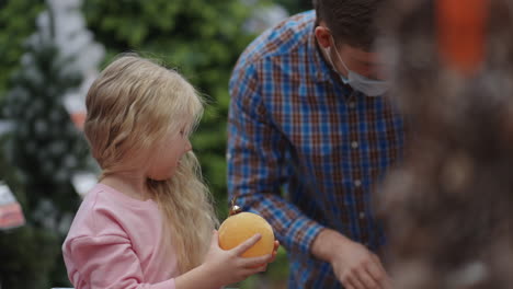 the girl holds a christmas ball in her hands in the store. a father in a medical mask on his face with his daughter choose jewelry and toys on christmas eve decorate the house and the christmas tree