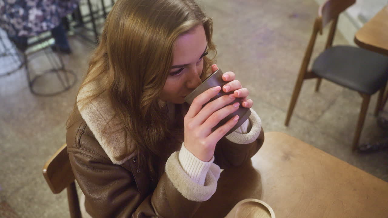 Up shot of woman sitting in cozy cafe sipping warm coffee, wearing brown shearling jacket, enjoying relaxed atmosphere with soft lighting, perfect for moments of tranquility and peaceful reflection
