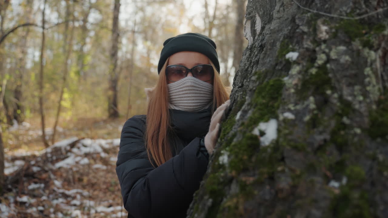 Mulher espreitadora se esconde atrás de uma árvore musgosa, espiando ao redor do tronco em uma floresta de outono nevada, óculos de sol e cachecol de malha escondem o rosto, gorro e jaqueta acolchoada indicam frio, e uma panorâmica cinematográfica lenta constrói o suspense.