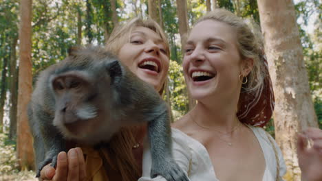 zwei frauen benutzen einen smartphone-video-chat und posieren mit einem affen, der auf der schulter sitzt. die besten freunde haben spaß mit affen und teilen abenteuer im tierpark. touristen reisen nach bali.