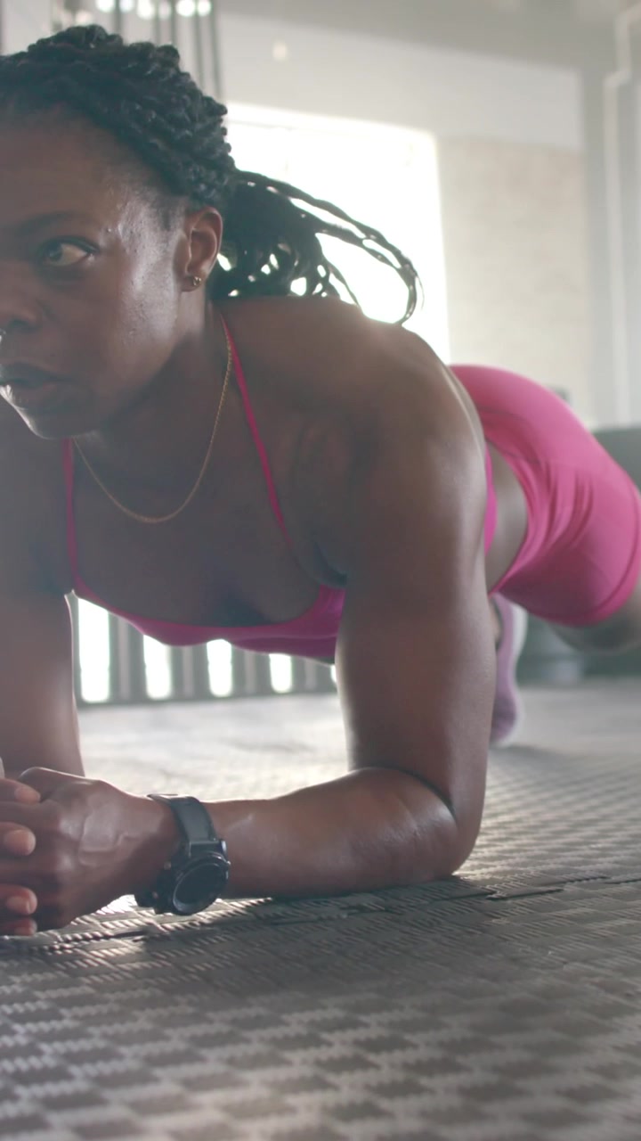 Vídeo vertical: Mulher afro-americana fazendo prancha em tapete de borracha com conjunto rosa e relógio de pulso.
