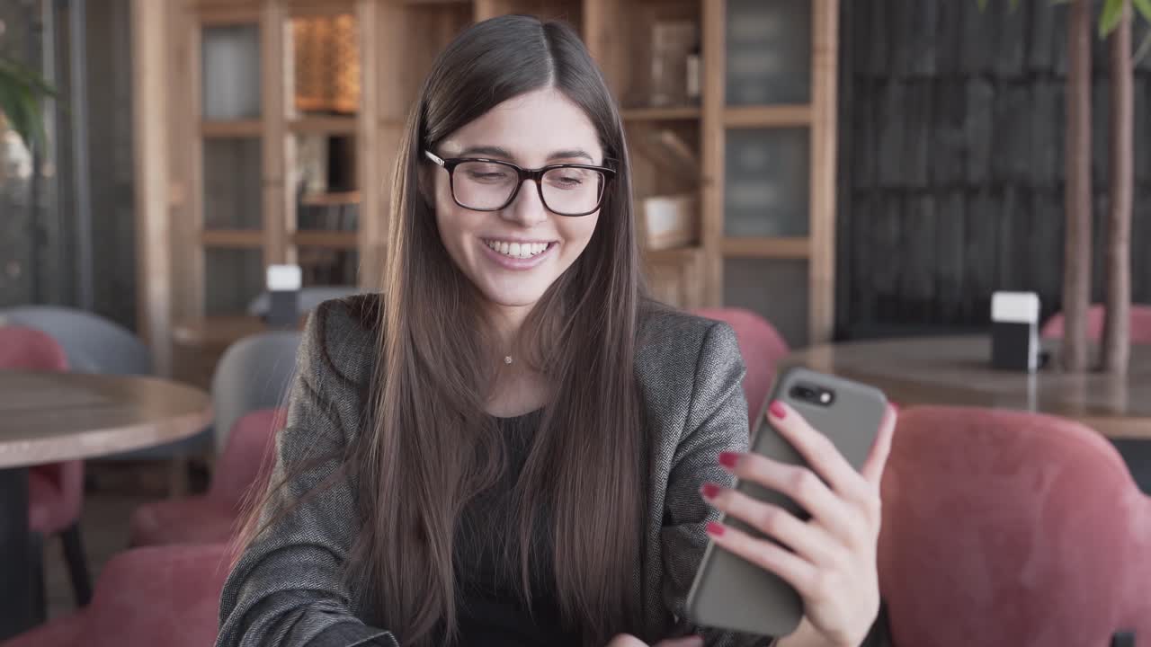 una joven guapa con una magnífica sonrisa con gafas, habla por teléfono inteligente en un acogedor café moderno y da un beso al aire