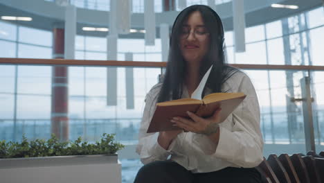 young girl with long black hair and glasses dances gently to music while reading book, seated indoors in modern mall area with glass railings, natural light, and elegant architecture surrounding her