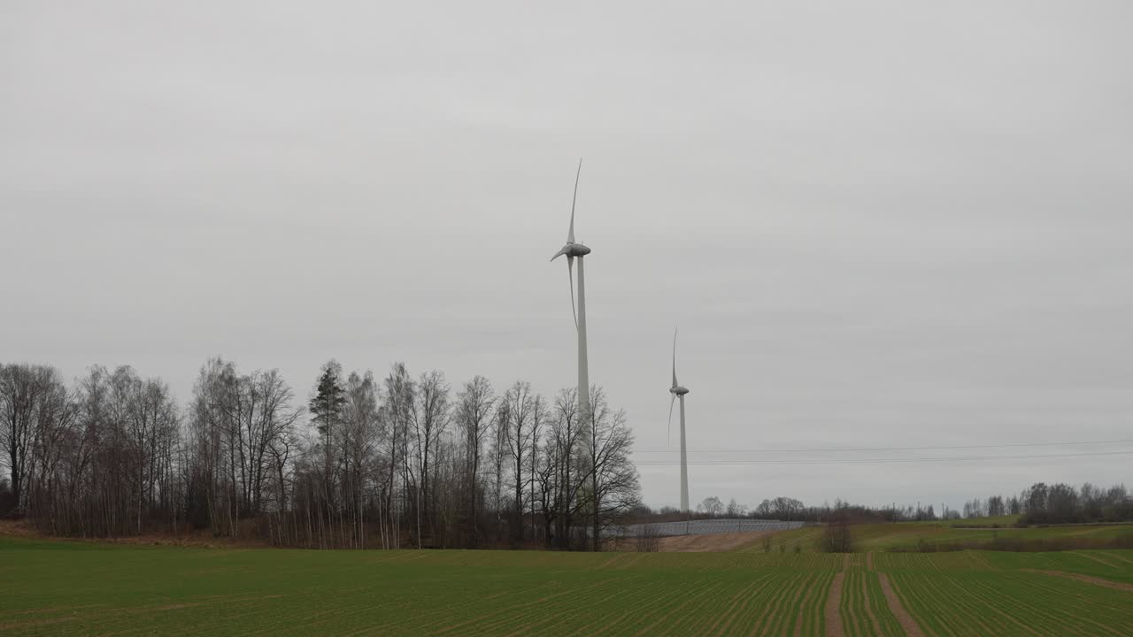 Vista panoramica di turbine eoliche in un tranquillo paesaggio rurale. Il cielo nuvoloso aggiunge un'atmosfera calma alla scena, mettendo in risalto i campi verdi e gli alberi in lontananza. Scatto grandangolare