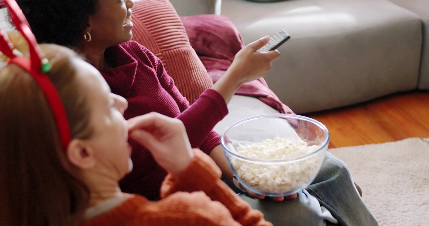 Forskellige kvinder derhjemme, klædt i bordeaux, trykker på fjernbetjeningen til tv'et, mens en ven tager en skål popcorn.