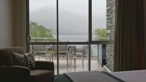 rainy day view from hotel room showing balcony furniture, misty lake, and mountain backdrop through large window from a side angle