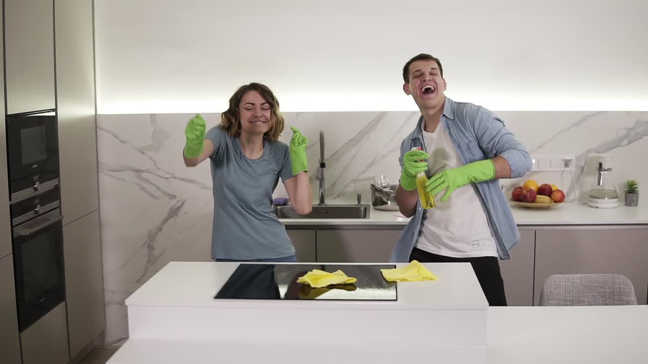 pareja feliz bailando en la cocina tanto en guantes de goma verdes divirtiéndose en el día de limpieza en el apartamento estudio. juventud moderna, la gente y el concepto de limpieza