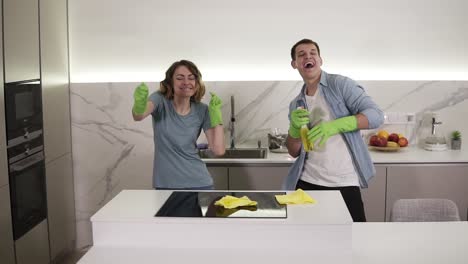 Happy-young-couple-dancing-in-kitchen-both-in-green-rubber-gloves-having-fun-on-clean-up-day-in-studio-apartment.-Modern-youth,-people-and-housekeeping-concept