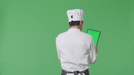 vista trasera del chef asiático usando una tableta mientras camina en el estudio de fondo de la pantalla verde