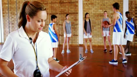 Female-coach-writing-on-clipboard-while-students-playing-in-basketball-court