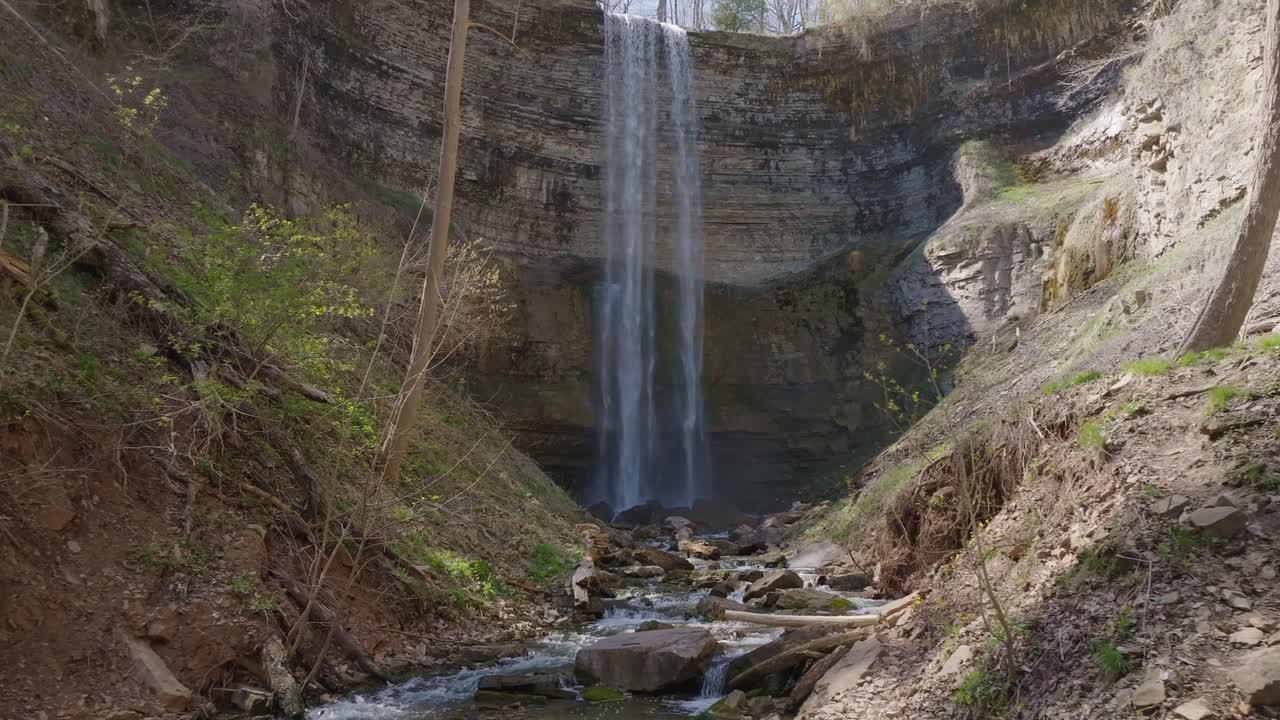 Høy foss som kaskader ned i en steinete bekk, omgitt av frodig grønt og jordfargede klipper.