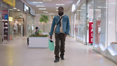 male shopper walking down bright mall corridor carrying green shopping bag over shoulder wearing denim jacket and hoodie with shoulder strap passing planters seating and store windows with sale signs