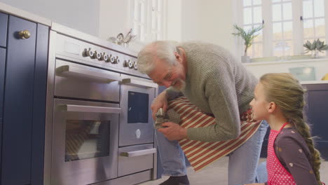 Grandfather-And-Granddaughter-Take-Freshly-Baked-Cupcakes-Out-Of-The-Oven-In-Kitchen-At-Home