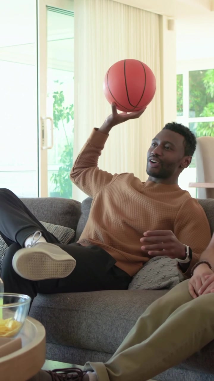 Video verticale: Uomo appoggiato sul divano di casa che passa una palla da basket, una chat inizia, indossa un maglione.