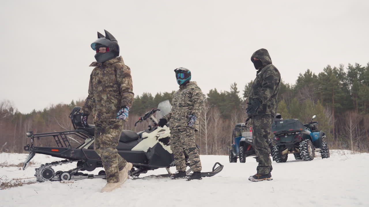 Squadra di motoslitte e ATV che si prepara in una radura innevata, piloti in attrezzatura mimetica controllano caschi e guanti, motoslitta parcheggiata accanto all'ATV, sfondo di pineta, cielo nuvoloso, tranquilla attesa.