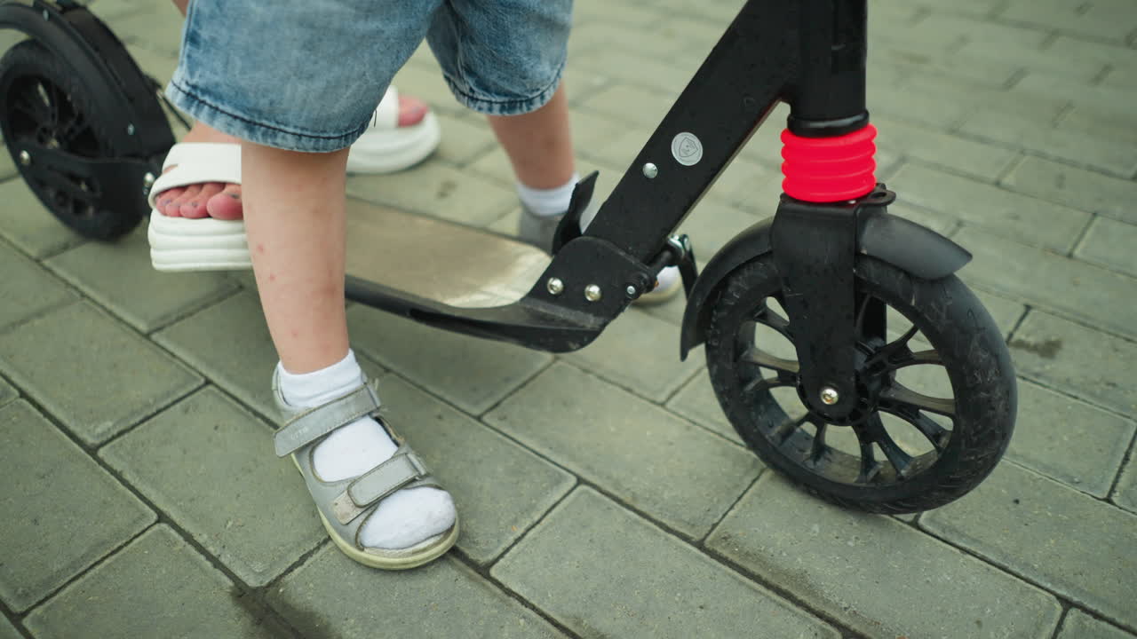 Leg view of a child and a woman standing on a black scooter. The child climbs the scooter as it slightly moves backward, the woman has one foot on the ground and another on the scooter