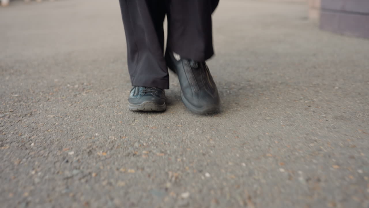 정면 뷰에서 카메라를 향해 걸어오는 검은색 가죽 신발을 신은 사람을 포착했어요. 낮은 앵글은 트레드, 움직임, 꾸준한 리듬을 강조하고, 바지 밑단은 햇빛 아래 땅을 스치며 목적 있는 도시의 움직임을 암시해요.