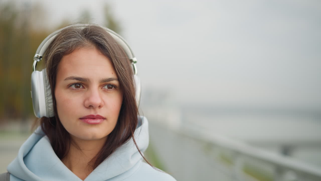 Bella ragazza che cammina maestosamente, guardando di lato mentre ascolta musica con le cuffie, con un'espressione sicura, in un ambiente esterno sereno e un contesto urbano tranquillo, con uno stile alla moda.