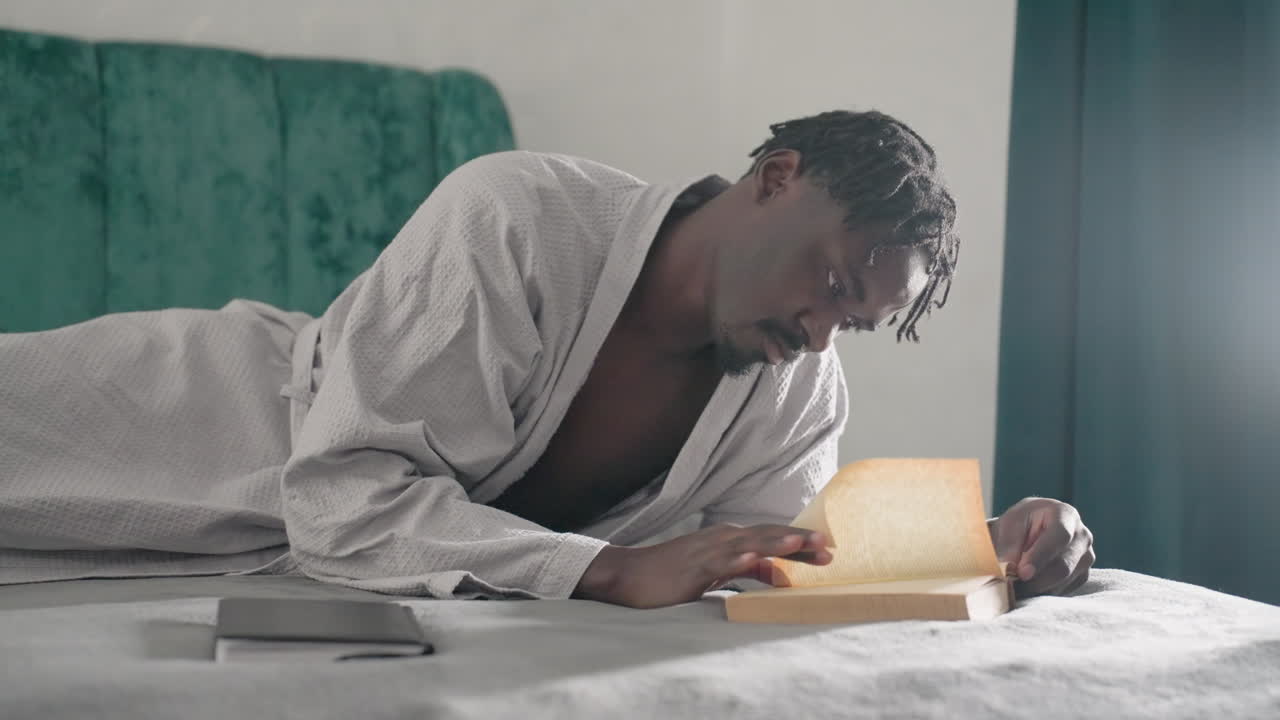 Man in white bathrobe laying on bed, focused on reading book with one hand, soft light highlighting book and fingers, green velvet headboard in background, peaceful indoor setting