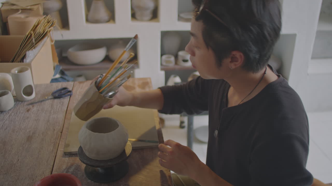 Asian Ceramist Choosing Brush for Decorating Vase in Pottery Workshop