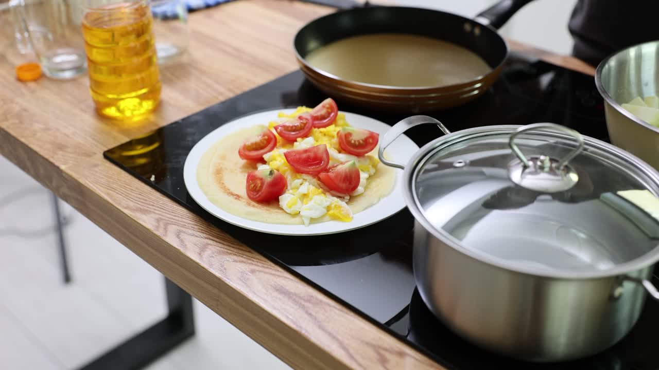 Breakfast preparation with pancake, scrambled eggs, and tomatoes in a modern kitchen