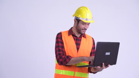 feliz hombre persa barbudo y guapo trabajador de la construcción con computadora portátil