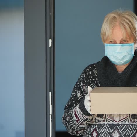 an elderly masked woman stands on the doorstep of the house and holds a small parcel in her hands